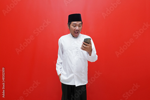 Asian muslim man with surprised expression looking at smartphone isolated on red background photo