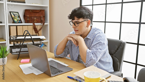 A thoughtful young man with glasses sits at a wooden desk, working on a laptop in a well-lit office.