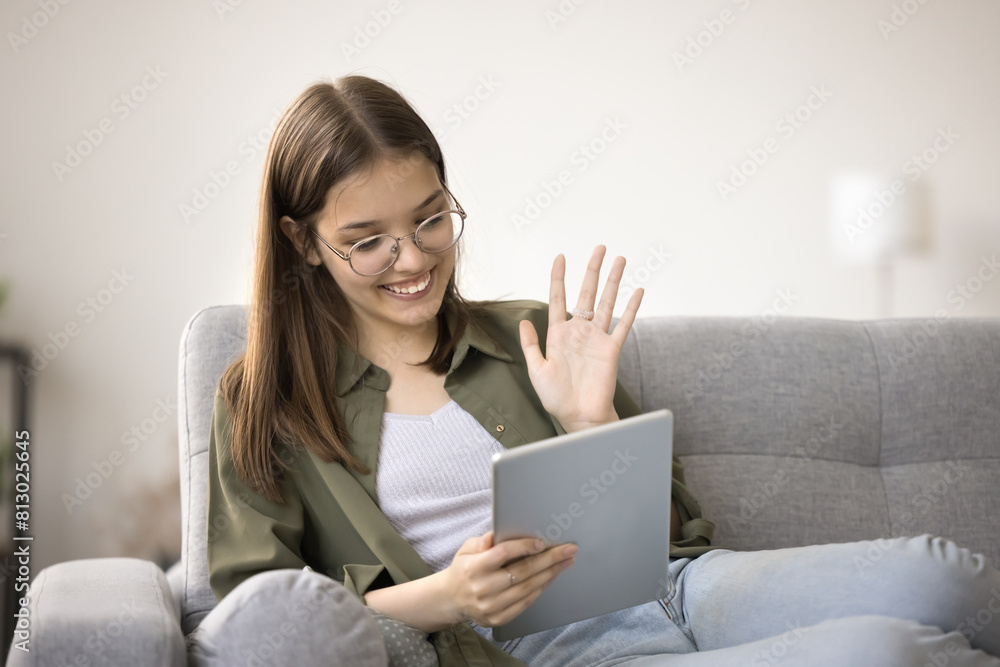 Happy pretty teenager girl in stylish eyeglasses saying hello on online video conference call, using tablet for remote Internet communication, waving greeting hand hello, smiling, laughing Happy pretty teenager girl in stylish eyeglasses saying hello on online video conference call, using tablet for remote Internet communication, waving greeting hand hello, smiling, laughing
