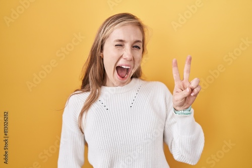 Young caucasian woman wearing white sweater over yellow background smiling with happy face winking at the camera doing victory sign with fingers. number two.