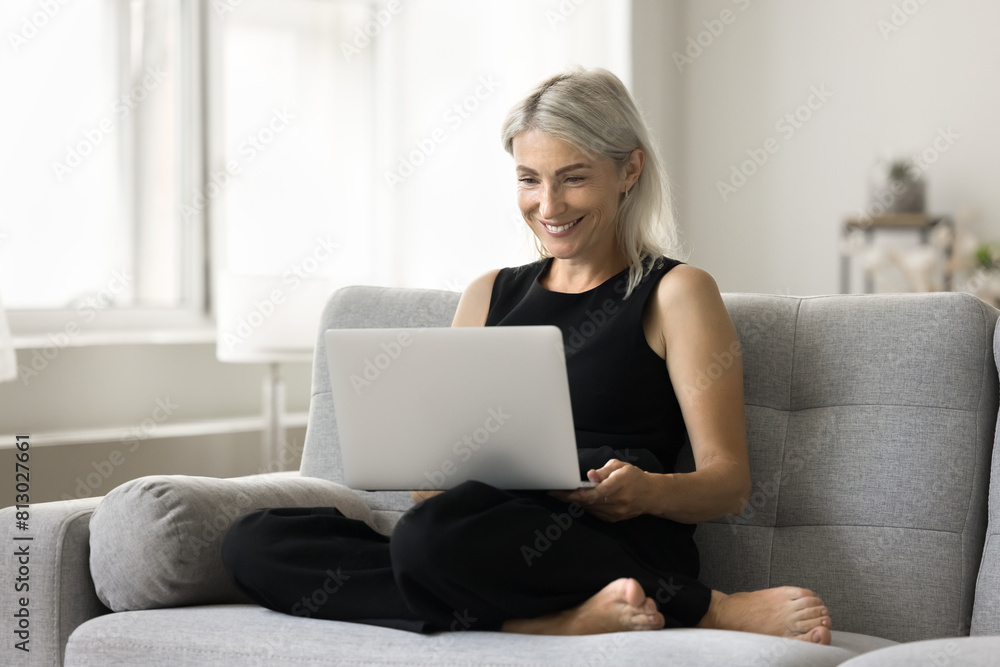 Fototapeta premium Happy mature freelancer woman sitting with gadget on home sofa, holding laptop computer on lap, using Internet technology for work communication, looking at screen, smiling, laughing