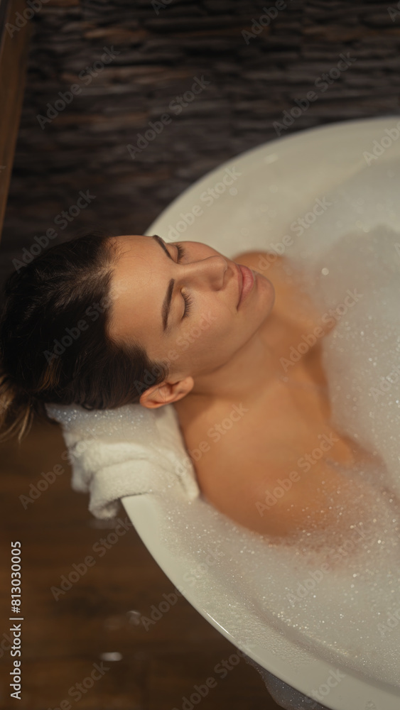 A serene young woman enjoys a relaxing bubble bath in a cozy, wood-accented bathroom. A serene young woman enjoys a relaxing bubble bath in a cozy, wood-accented bathroom.
