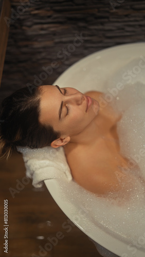A serene young woman enjoys a relaxing bubble bath in a cozy  wood-accented bathroom.