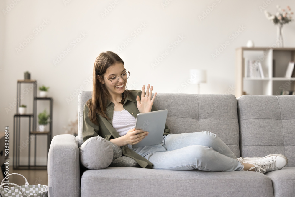 Positive smart teenage school student girl talking on video call chat on tablet, relaxing on home sofa, waving greeting hand hello, saying hi, smiling, laughing, enjoying Internet communication Positive smart teenage school student girl talking on video call chat on tablet, relaxing on home sofa, waving greeting hand hello, saying hi, smiling, laughing, enjoying Internet communication