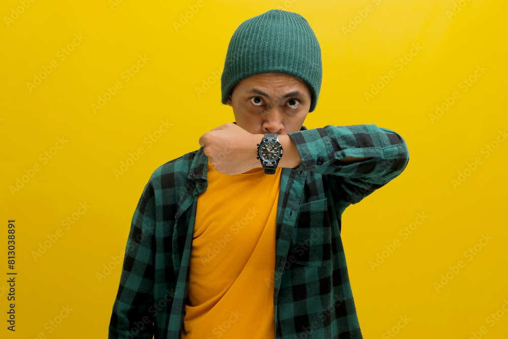 Impatient Asian man in a beanie and casual shirt shows his watch to the camera, reminding about a schedule, target, or deadline. Isolated on a yellow background Impatient Asian man in a beanie and casual shirt shows his watch to the camera, reminding about a schedule, target, or deadline. Isolated on a yellow background