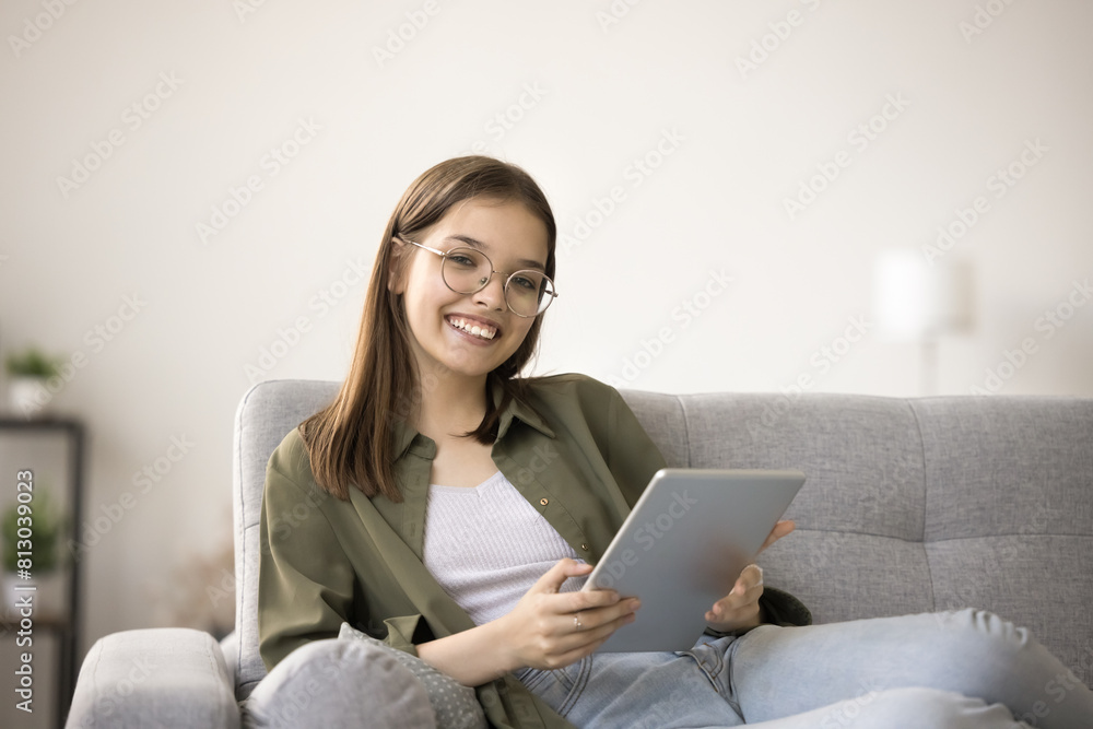 Cheerful pretty teen schoolgirl in eyeglasses doing school homework on tablet, holding gadget, resting on couch at home, using digital gadget, modern technology for education, looking at camera Cheerful pretty teen schoolgirl in eyeglasses doing school homework on tablet, holding gadget, resting on couch at home, using digital gadget, modern technology for education, looking at camera