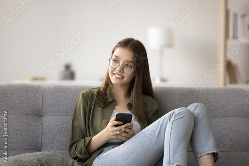 Positive dreamy pretty teen girl in glasses holding smartphone, looking away with toothy smile, thinking, dreaming, relaxing on sofa, enjoying domestic Internet communication, wireless technology