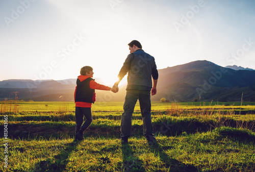 Father and son walking through picturesque mountain valley at sunset holding hands, back view