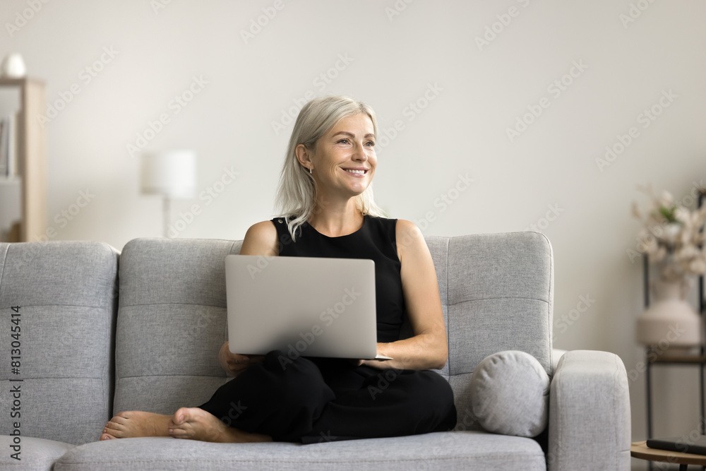 Cheerful thoughtful senior woman holding laptop on lap, sitting on couch, looking away, thinking, dreaming, planning retirement, using wireless Internet technology, communication Cheerful thoughtful senior woman holding laptop on lap, sitting on couch, looking away, thinking, dreaming, planning retirement, using wireless Internet technology, communication