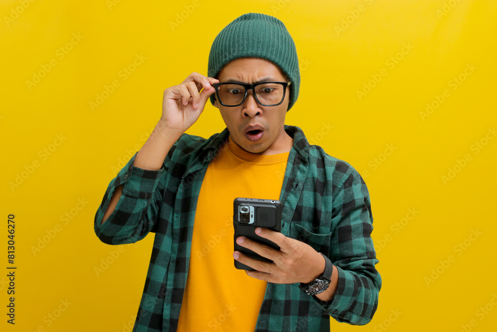 Shocked Asian man, sporting a beanie hat and casual shirt, adjusts his glasses while gazing at his smartphone with a surprised expression, exclaiming Wow, Great News Standing against yellow background Shocked Asian man, sporting a beanie hat and casual shirt, adjusts his glasses while gazing at his smartphone with a surprised expression, exclaiming Wow, Great News Standing against yellow background