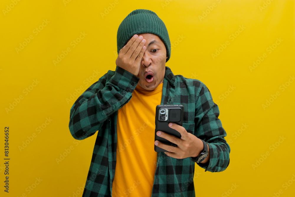 Shocked Asian man, dressed in a beanie hat and casual shirt, covers his eyes in a comical and surprised reaction while looking at his phone, clearly taken aback by unexpected news, yellow background Shocked Asian man, dressed in a beanie hat and casual shirt, covers his eyes in a comical and surprised reaction while looking at his phone, clearly taken aback by unexpected news, yellow background