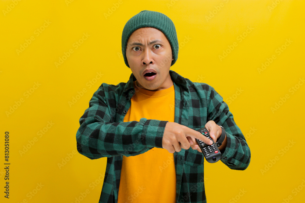 Surprised young Asian man, clad in a beanie hat and casual shirt, watches shocking news on TV while holding a remote, expressing a mix of shock, confusion, and fear, isolated on a yellow background Surprised young Asian man, clad in a beanie hat and casual shirt, watches shocking news on TV while holding a remote, expressing a mix of shock, confusion, and fear, isolated on a yellow background