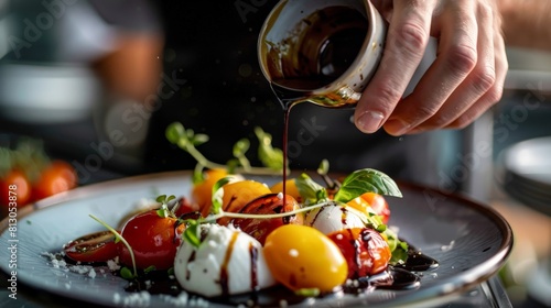 A chef is carefully pouring a balsamic vinegar reduction over a composed salad of heirloom tomatoes, creamy burrata cheese, and fresh basil.
