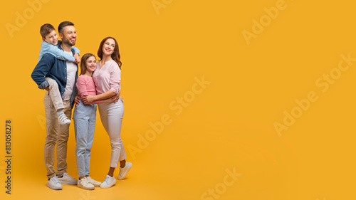 A happy family consisting of a father, mother, son, and daughter are standing closely together, embracing with smiles on their faces, looking at copy space on yellow background