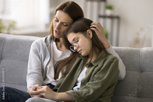Compassionate young mother comforting depressed sad teen girl, hugging daughter with love, care, speaking, giving sympathy, advice, support, holding kids hand, sitting on home couch