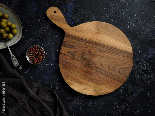 Top view of empty round wooden cutting board with handle, red pepper and green olives on abstract dark background. photo