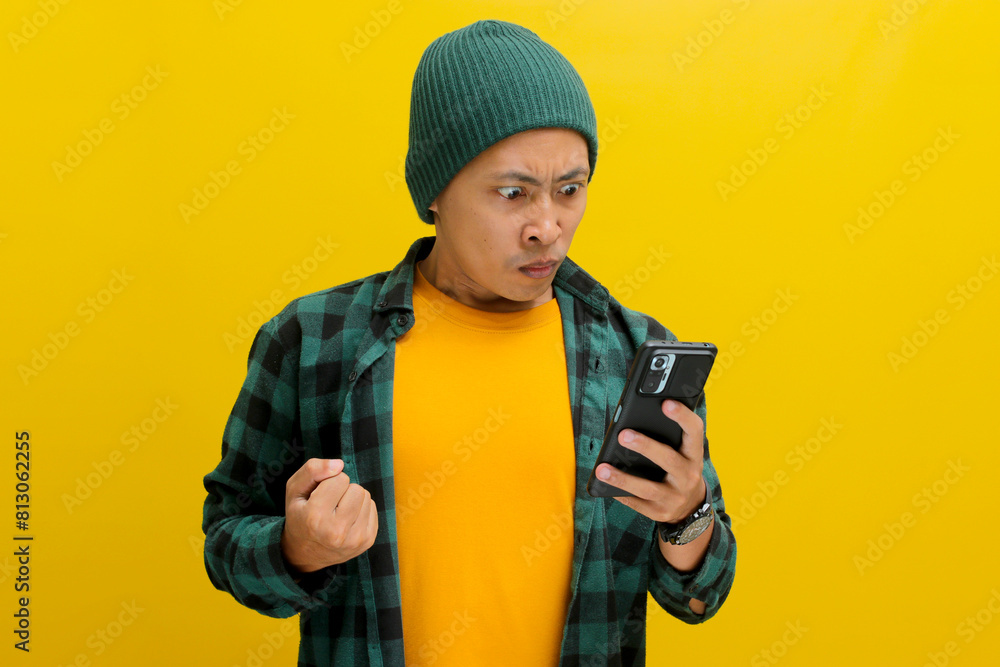 An angry Asian man, dressed in a beanie hat and casual shirt, visibly reacts to bad news while looking at his mobile phone, while standing against yellow background An angry Asian man, dressed in a beanie hat and casual shirt, visibly reacts to bad news while looking at his mobile phone, while standing against yellow background
