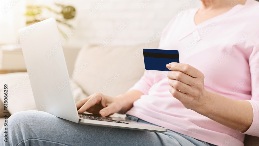 Naklejka premium Cropped of woman is seated on a couch, holding a credit card in one hand and a laptop in the other. She appears focused on the laptop screen while balancing the credit card in her hand.