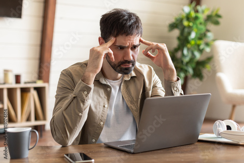 Fotografie A bearded man is seated at a wooden table with a laptop, exuding an expression of concern and deep concentration