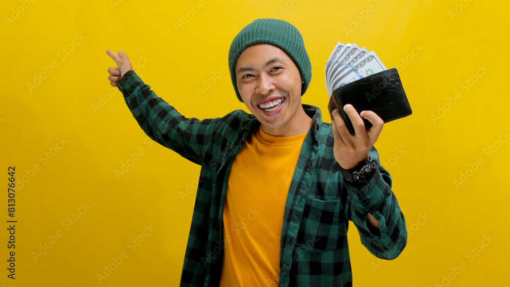 An enthusiastic young Asian man, dressed in a beanie hat and casual shirt, points to an empty space for text or advertisement while holding a wallet, isolated on a yellow background An enthusiastic young Asian man, dressed in a beanie hat and casual shirt, points to an empty space for text or advertisement while holding a wallet, isolated on a yellow background
