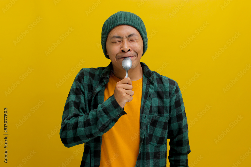 Excited, hungry young Asian man, donning a beanie hat and casual shirt, envisions delicious food, holding a spoon near his mouth with an excited and joyful expression, isolated on yellow background Excited, hungry young Asian man, donning a beanie hat and casual shirt, envisions delicious food, holding a spoon near his mouth with an excited and joyful expression, isolated on yellow background