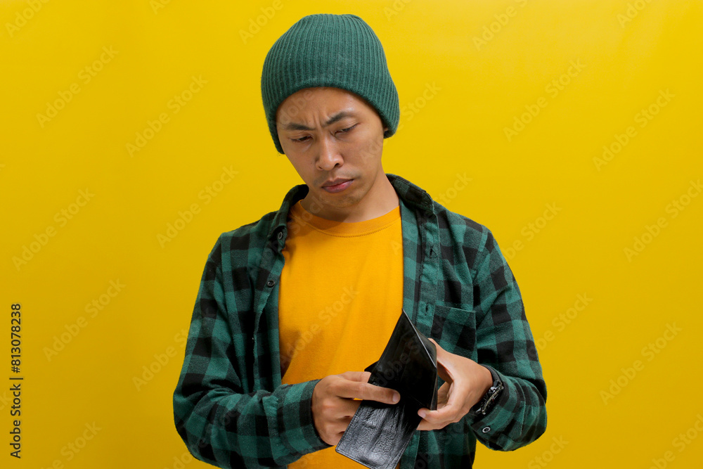 Frowning young Indonesian man holds an empty wallet, isolated on a yellow background Frowning young Indonesian man holds an empty wallet, isolated on a yellow background