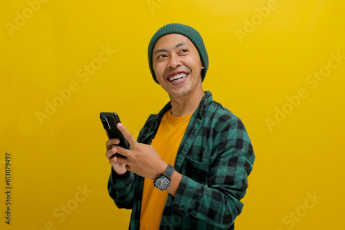 Happy and amazed Asian man, sporting a beanie hat and casual shirt, is using his smartphone for texting, social media engagement, online ecommerce shopping in mobile apps, or watching videos