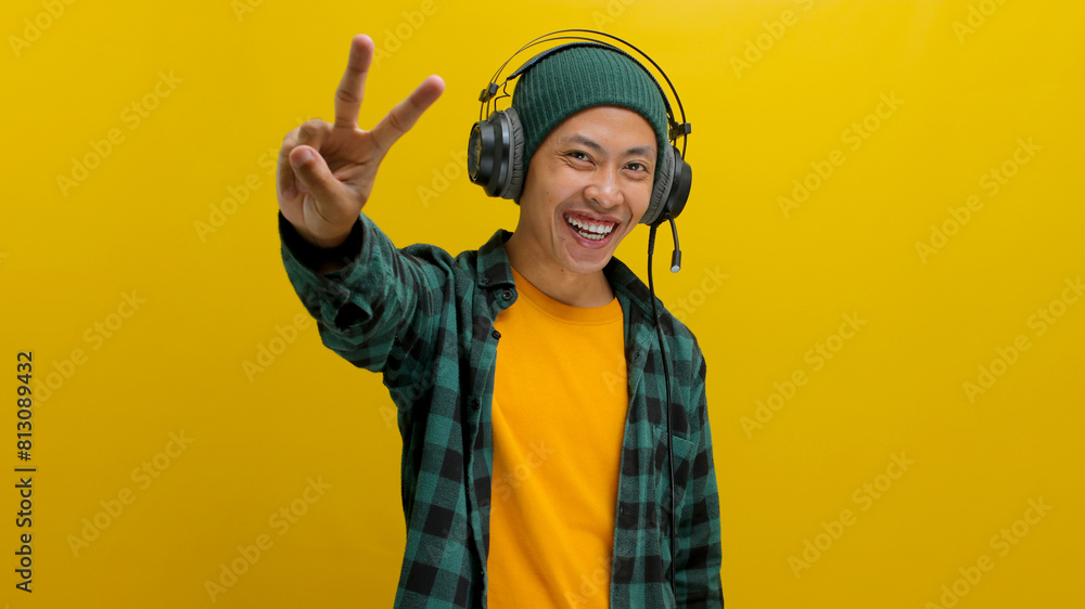 Smiling Asian man in a beanie and casual clothes throws up a peace sign while listening to music or a podcast on his headphones. Isolated on a yellow background. Smiling Asian man in a beanie and casual clothes throws up a peace sign while listening to music or a podcast on his headphones. Isolated on a yellow background.