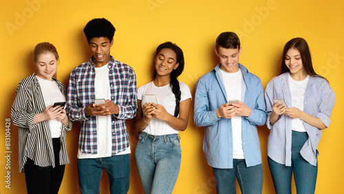 A diverse group of five multiethnic teenagers stand side-by-side against a vibrant yellow backdrop, each engrossed in their own smartphone