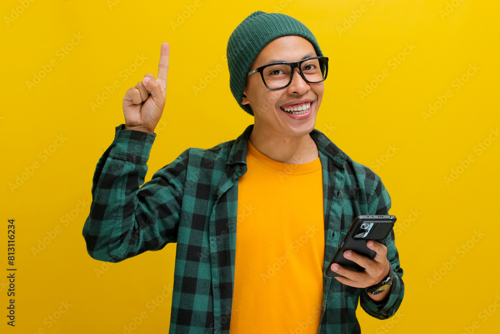 Asian man wearing beanie hat, casual shirt, and eyeglasses points at an empty space while confidently holding his phone, displaying excitement, happiness, and confidence, isolated on yellow background Asian man wearing beanie hat, casual shirt, and eyeglasses points at an empty space while confidently holding his phone, displaying excitement, happiness, and confidence, isolated on yellow background