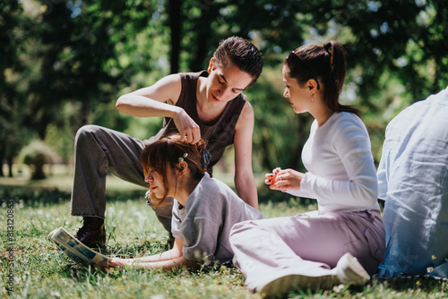 Three young friends enjoy a leisurely day in the park, with one braiding another's hair while sitting on the grass.