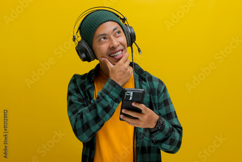 Young Asian man, deep in thought while listening to music on headphones. He holds his phone and gazes upwards, seemingly lost in the music or browsing the music app. Isolated on a yellow background.
