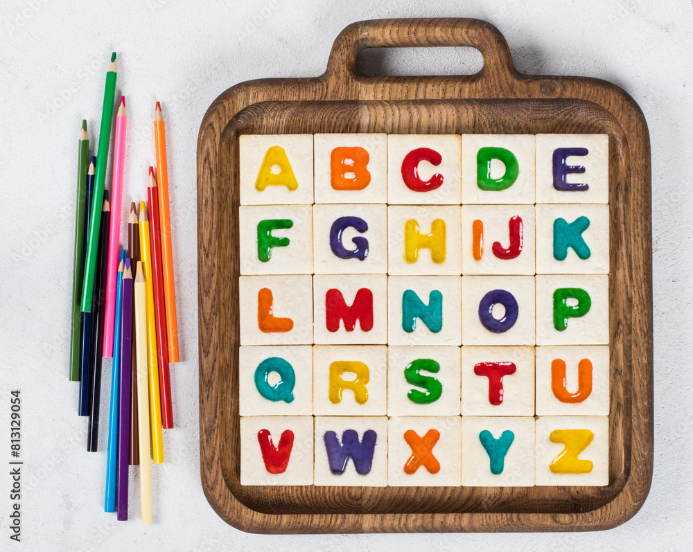 Children Cookies English Alphabet. Square cookies with colored marmalade filling in the shape of English letters. On a wooden plate. With colored pencils. White background. Top view. Children Cookies English Alphabet. Square cookies with colored marmalade filling in the shape of English letters. On a wooden plate. With colored pencils. White background. Top view.