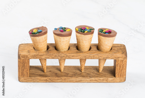 Candy mini Waffle cone, with chocolate nut cream and colored dragees. On a wooden serving stand. White background