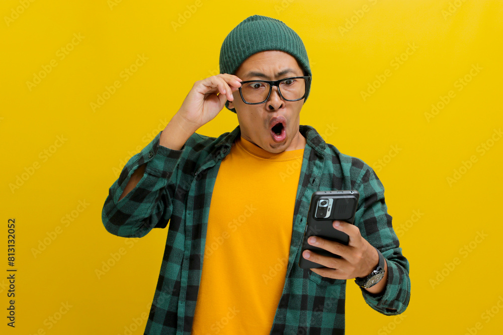 Shocked Asian man, sporting a beanie hat and casual shirt, adjusts his glasses while gazing at his smartphone with a surprised expression, exclaiming Wow, Great News Standing against yellow background Shocked Asian man, sporting a beanie hat and casual shirt, adjusts his glasses while gazing at his smartphone with a surprised expression, exclaiming Wow, Great News Standing against yellow background