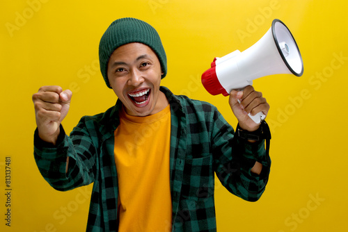 Happy and successful Asian man in a beanie hat clenches his fist in a victory gesture while shouting and screaming loudly into a megaphone. Communication concept. Isolated on a yellow background photo