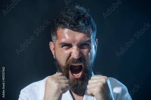 Angry screaming bearded man in white kimono in fighting position. Karate fighter preparing for competition. Muscular male fitness model ready for karate training. Strong martial arts master on fight.