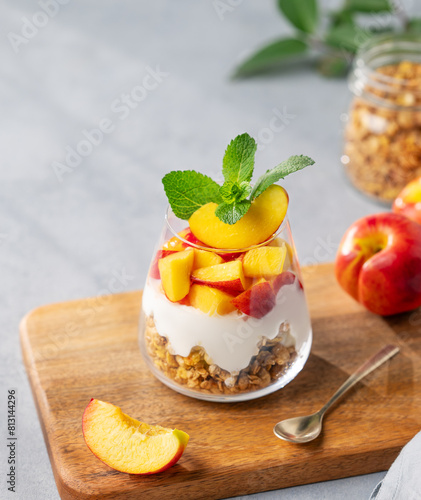 Greek yogurt parfait with peach and granola in a glass on a wooden board on a light background with fresh fruits.