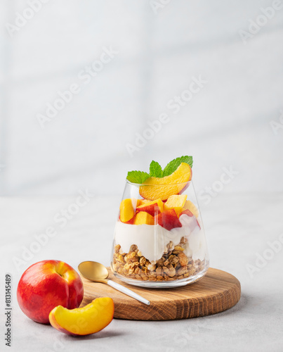 Yogurt parfait with peach and muesli in a glass on a wooden board on a light background with fresh fruits and shadow. Healthy eating concept.