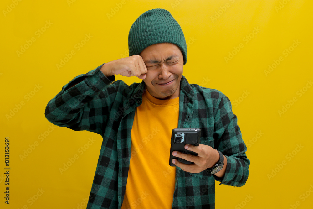 An unhappy Asian man, dressed in a beanie hat and casual shirt, holds a phone, visibly upset, displaying sadness, worry, or perhaps shedding tears in reaction to distressing news received online An unhappy Asian man, dressed in a beanie hat and casual shirt, holds a phone, visibly upset, displaying sadness, worry, or perhaps shedding tears in reaction to distressing news received online