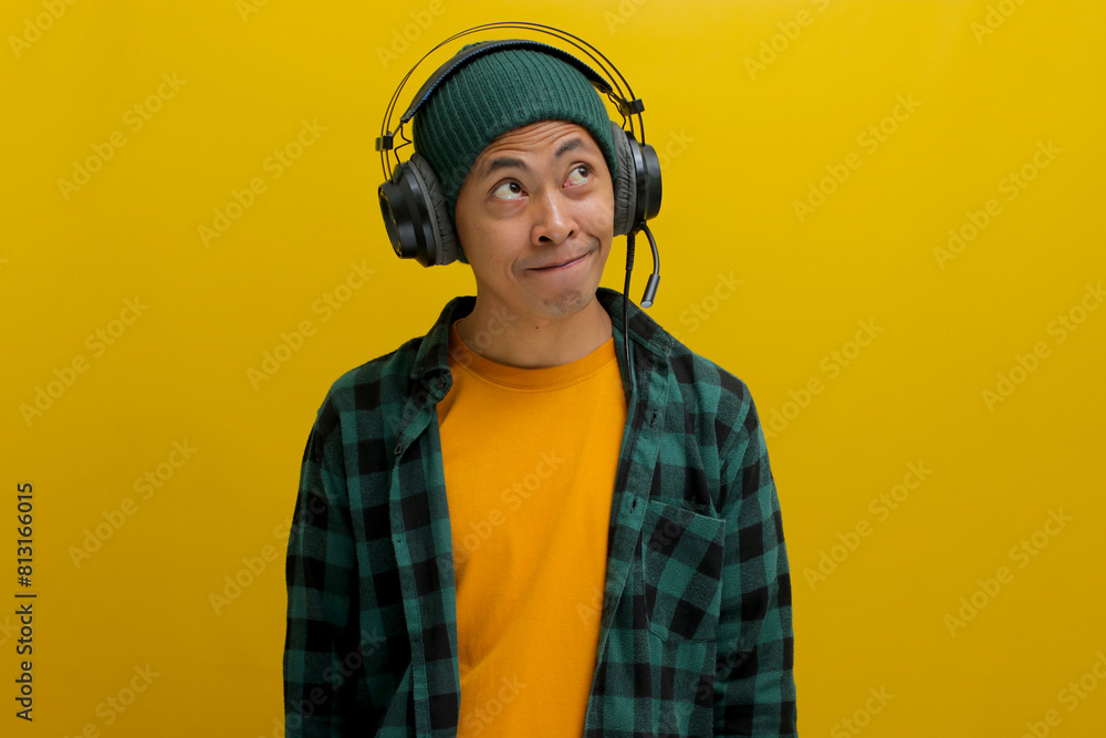 Young Asian man in a beanie and casual clothes, lost in thought while listening to music on his headphones. He gazes upward, a thoughtful smile playing on his lips. Isolated on a yellow background Young Asian man in a beanie and casual clothes, lost in thought while listening to music on his headphones. He gazes upward, a thoughtful smile playing on his lips. Isolated on a yellow background