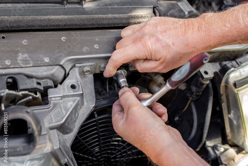 an auto mechanic fixes the car engine cooling fan under the radiator grill with a ratchet wrench. Car repair and maintenance. © Natalia