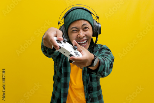 An enthusiastic Asian gamer, adorned in a beanie hat, casual shirt, and headphones, celebrates victory while playing a video game on a console with a gamepad or joystick isolated on yellow background.