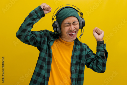 Young Asian man in a beanie and casual clothes dances with joy while listening to music on headphones. Isolated on a yellow background.