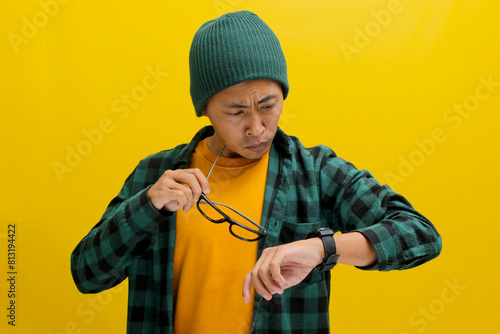 Annoyed Asian man glances at his watch, clearly frustrated by his friend's tardiness for their appointment. Isolated on a yellow background.