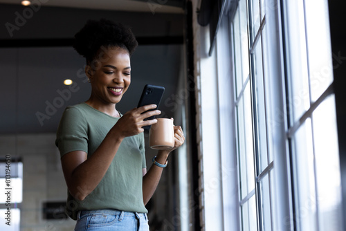 At office, African American businesswoman holding coffee, looking at phone, copy space