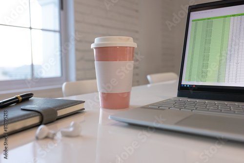At office, laptop displaying spreadsheet on white desk beside pink coffee cup photo