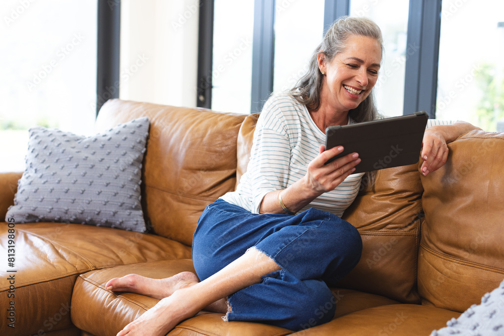 At home, mature Caucasian woman laughing, holding tablet on brown sofa At home, mature Caucasian woman laughing, holding tablet on brown sofa