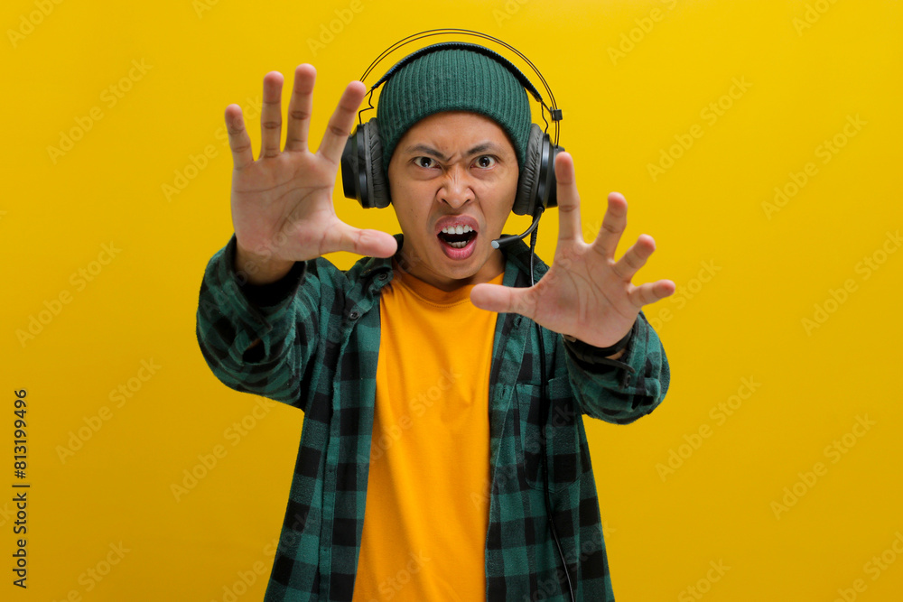 Annoyed Asian man in a beanie and casual clothes holds his hand up in a stop gesture (palm facing camera) while listening to music on headphones. Isolated on a yellow background. Annoyed Asian man in a beanie and casual clothes holds his hand up in a stop gesture (palm facing camera) while listening to music on headphones. Isolated on a yellow background.