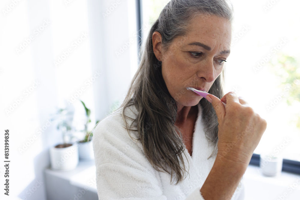 At home, mature Caucasian woman brushing teeth in a white robe At home, mature Caucasian woman brushing teeth in a white robe